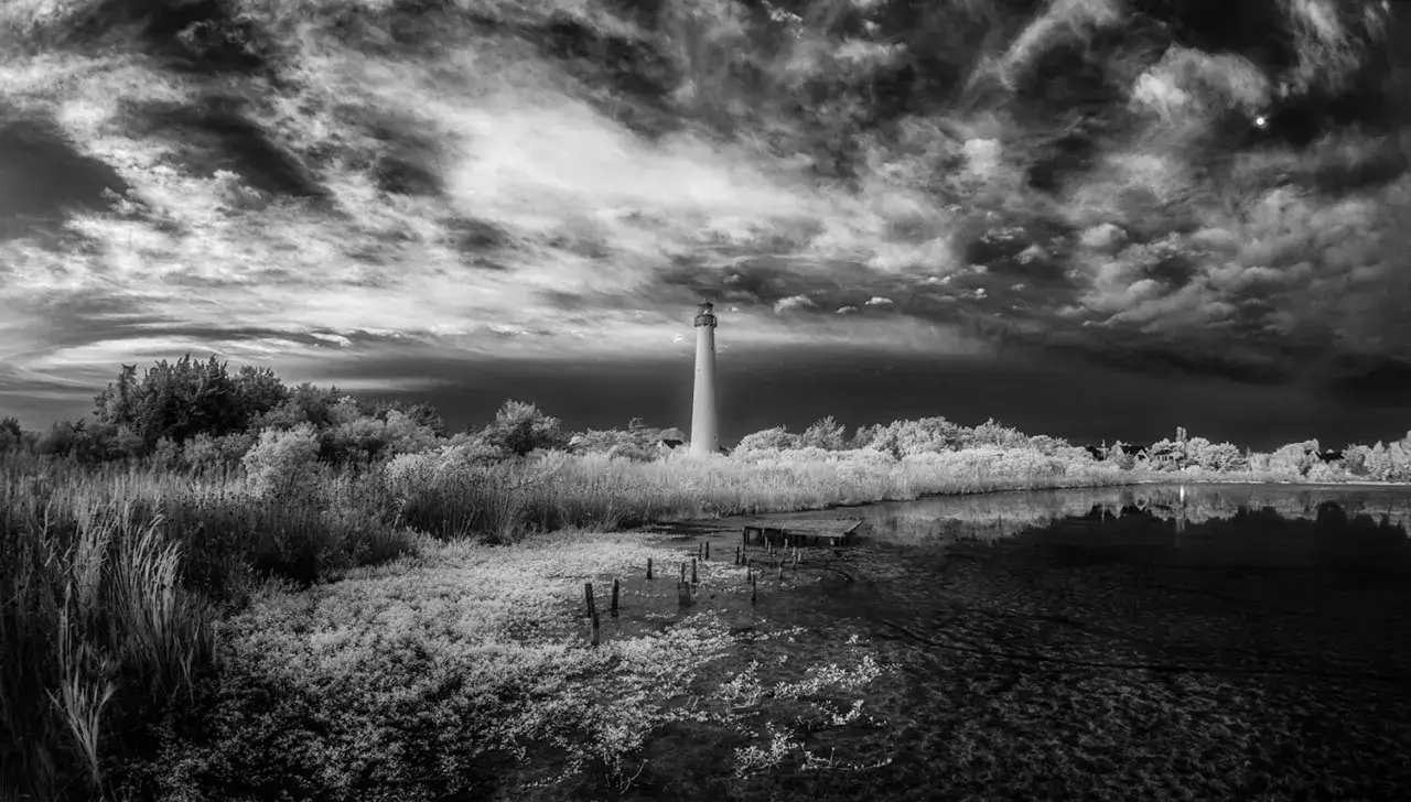 Faro blanco sobre un paisaje surrealista, con vegetaci&oacute;n brillante y un cielo dram&aacute;tico. Fotograf&iacute;a infrarroja que revela un mundo et&eacute;reo.